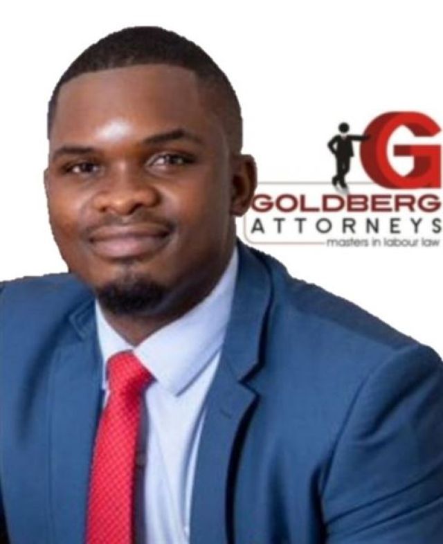 a black man dressed in a formal blue suit, light blue shirt, and red tie. He has short hair and a neatly trimmed beard and is smiling slightly while facing the camera. Behind him, there’s a logo that reads “GOLDBERG ATTORNEYS – masters in labour law”, featuring a stylized red G and a human figure incorporated into the design. The background is white, giving the photo a clean, professional appearance.